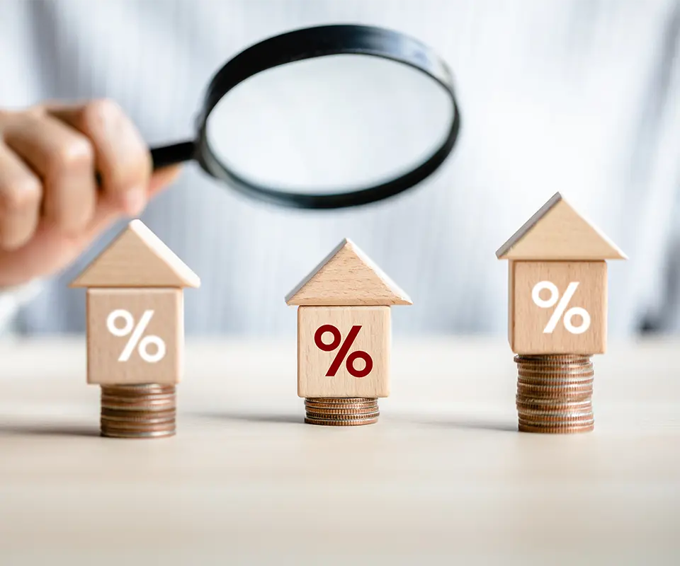 A man looking with a magnifying glass to three wooden houses and some coins.